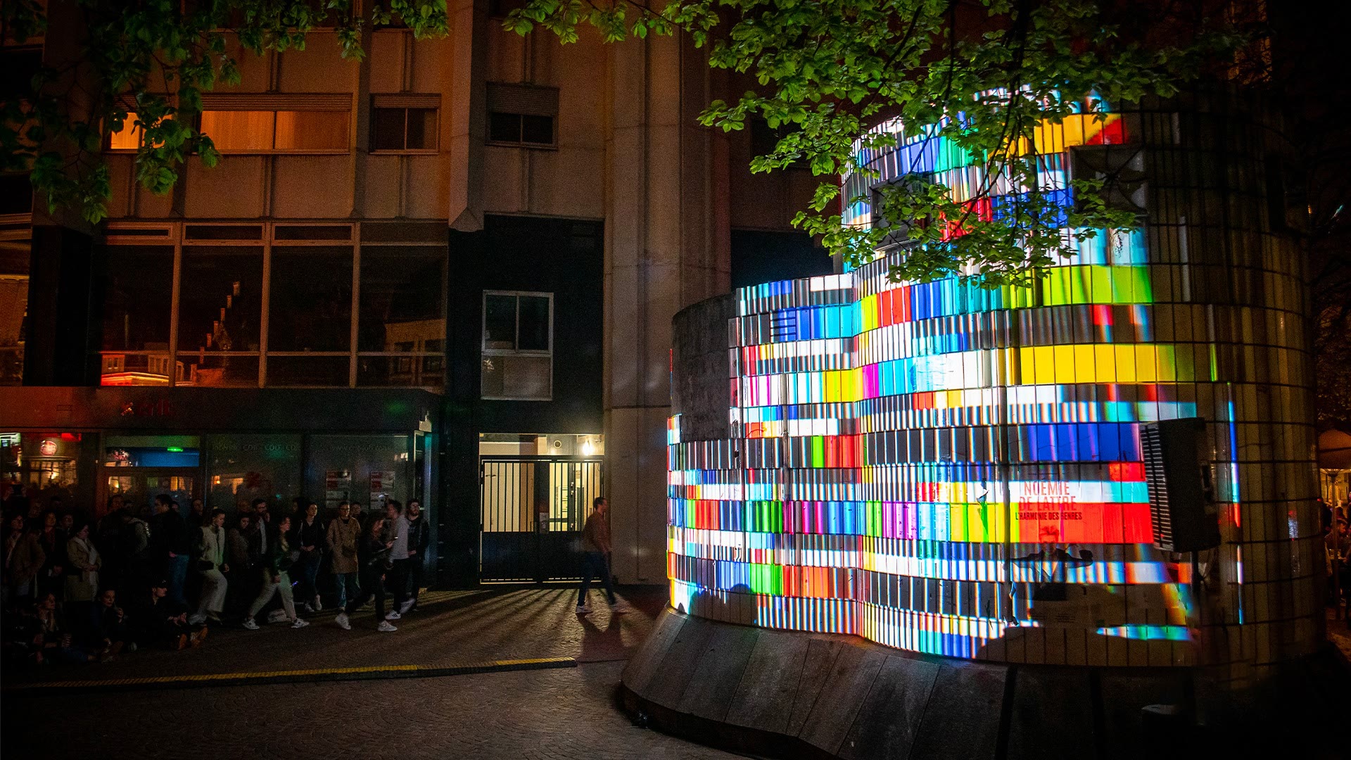 874 seen from the street at Nouveau Siècle in Lille, the curved concrete ventilation shaft covered in fine vertical stripes of saturated colour, the concrete building and a lit shopfront behind it and a group of people walking past on the pavement