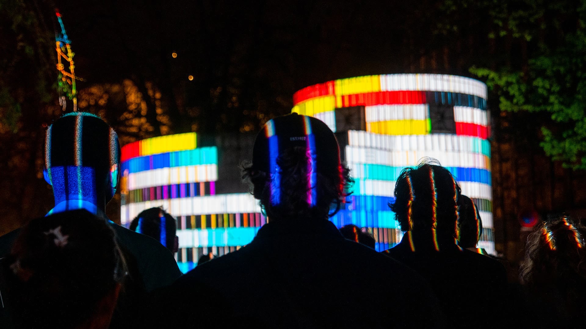 874 seen from within the audience, heads silhouetted across the foreground against the shaft mapped in hard blocks of red, yellow, cyan and white, dark trees closing in on either side