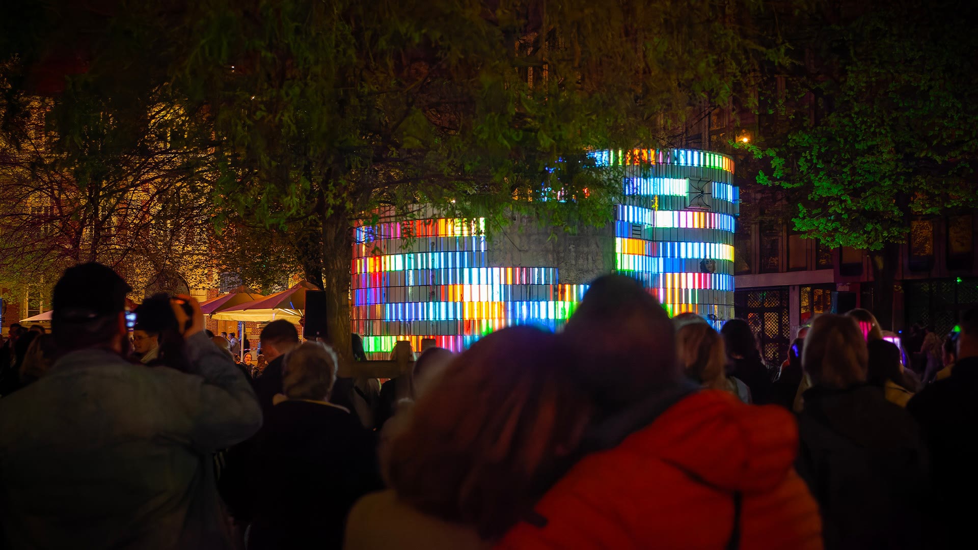 874 with a tree crossing the foreground, the shaft striped in dense multicoloured bands, visitors watching in the dark and a lit market stall to the left