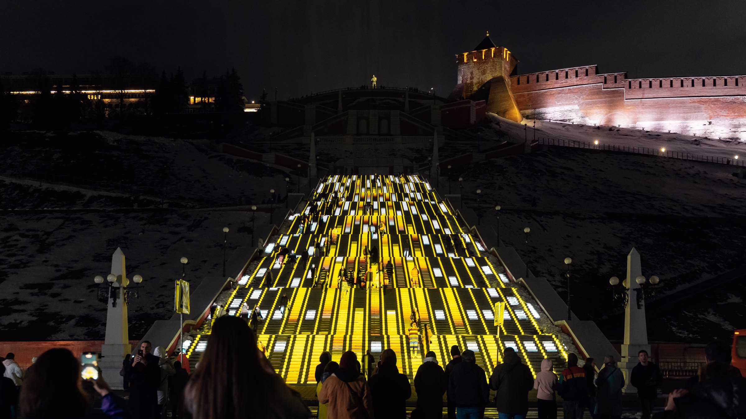 Ascent on the Chkalov Staircase seen head-on from the foot of the flight, gold and white bands of light stacked across every step, a crowd watching from the embankment below and the floodlit kremlin wall and tower on the hillside to the right