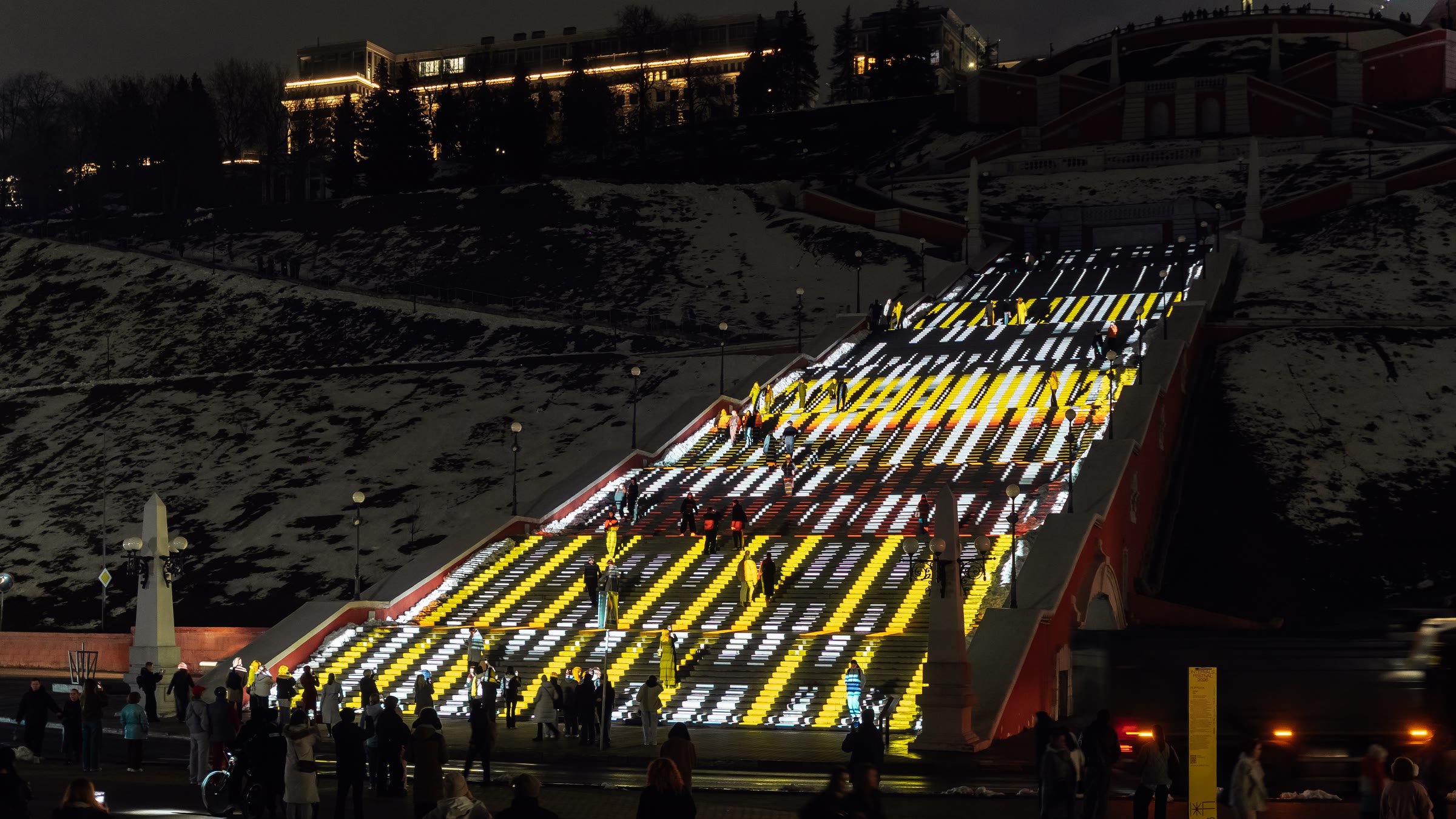 Ascent seen from the side, the full diagonal run of the Chkalov Staircase striped in yellow, white and red light, snow covering the dark hillside on either side and visitors gathered along the embankment at the foot of the steps