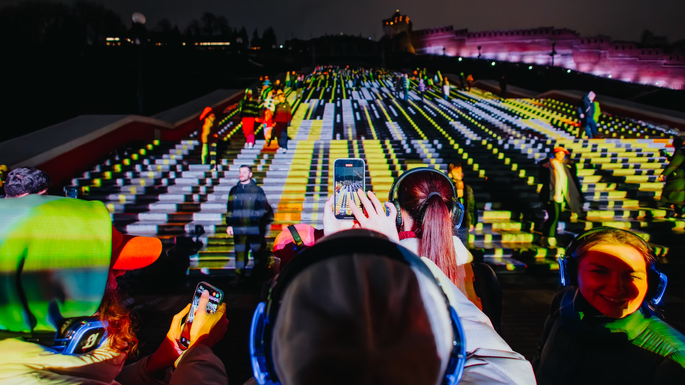 Ascent from inside the audience, visitors in lit headphones filming on their phones as yellow, white and green blocks of light run up the staircase ahead of them, the kremlin wall glowing pink along the ridge behind