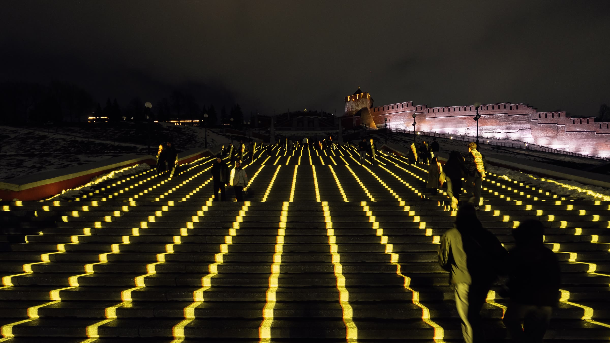 Ascent in a quieter passage, continuous gold lines running straight up the treads of the Chkalov Staircase in a radiating fan, a few figures climbing among them and the floodlit kremlin wall and tower on the ridge to the right